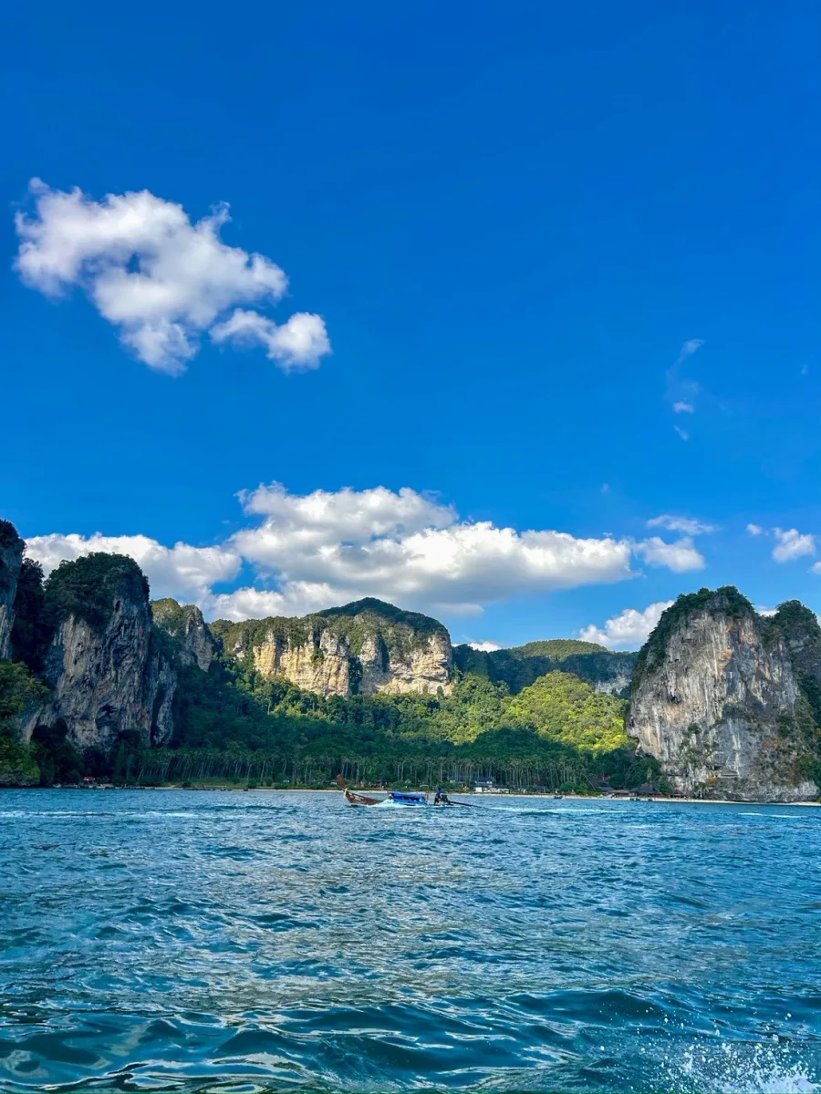 เรือหางยาวแล่นผ่านหน้าผาหินปูนและน้ำทะเลสีครามที่ไร่เลย์ กระบี่